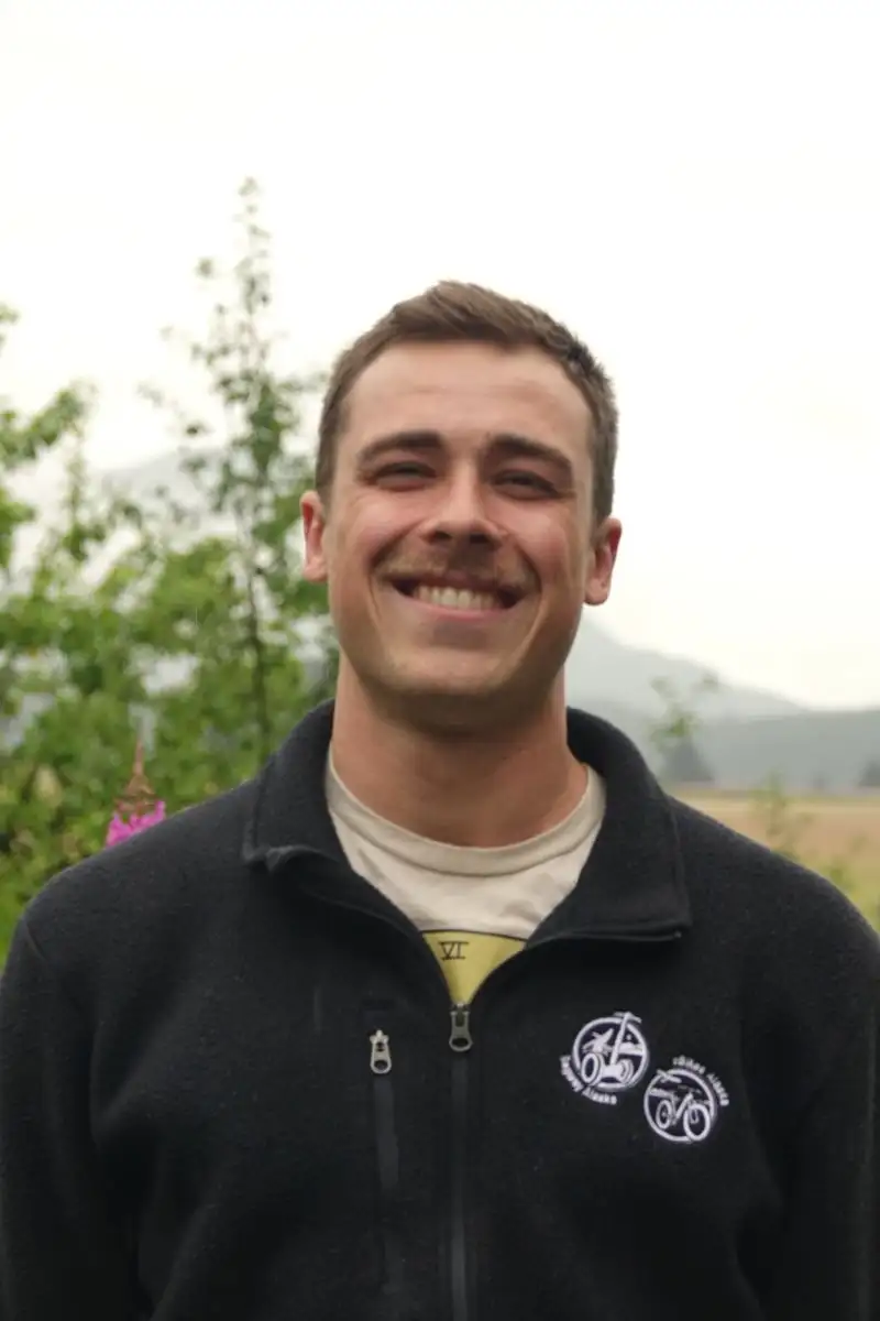 Get to Know Us 9 A smiling young man with a mustache wearing a black fleece jacket outdoors.