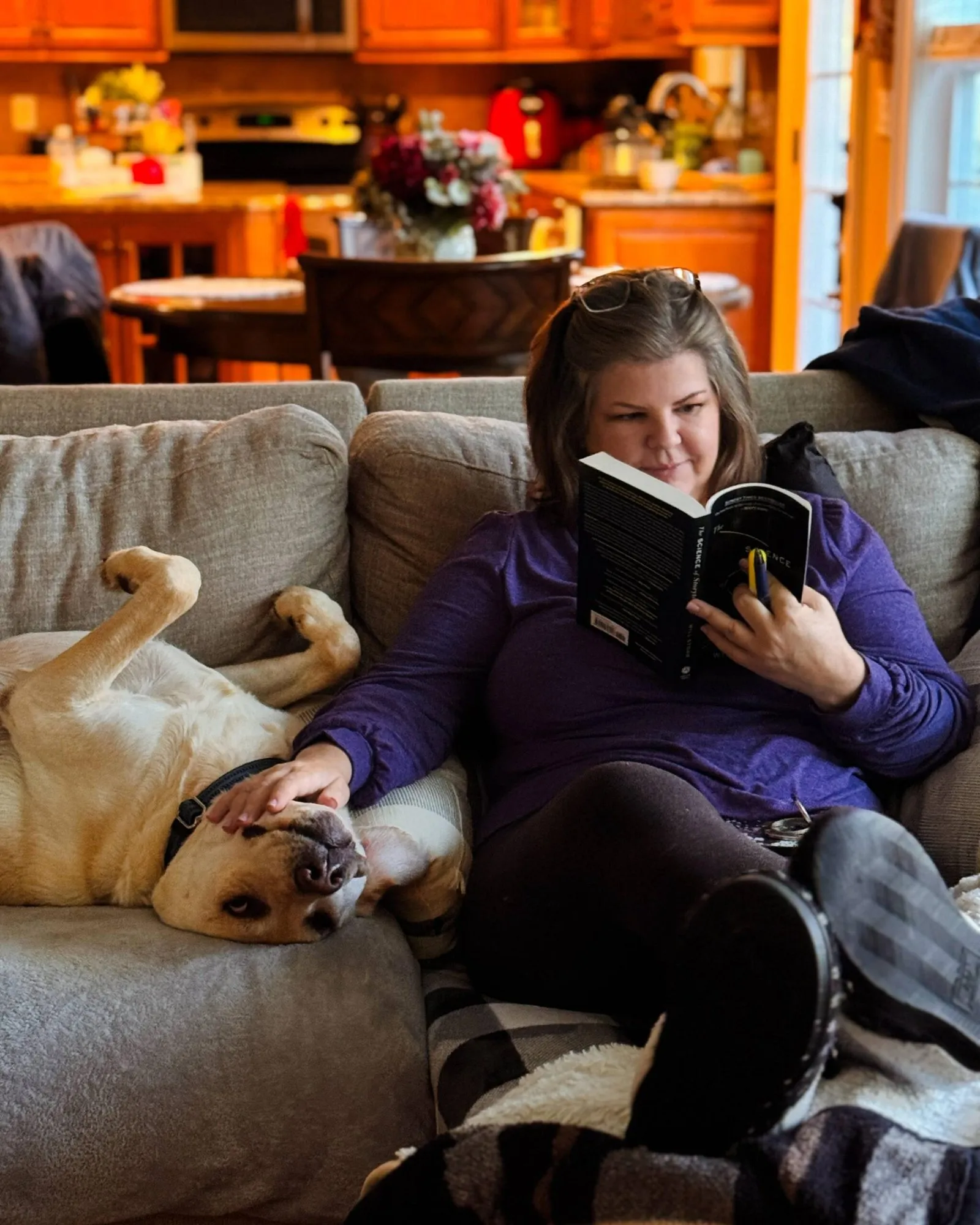 Jeannie Ruesch reading The Science of Storytelling on the couch, one hand resting on her yellow Labrador, who is sprawled upside down beside her