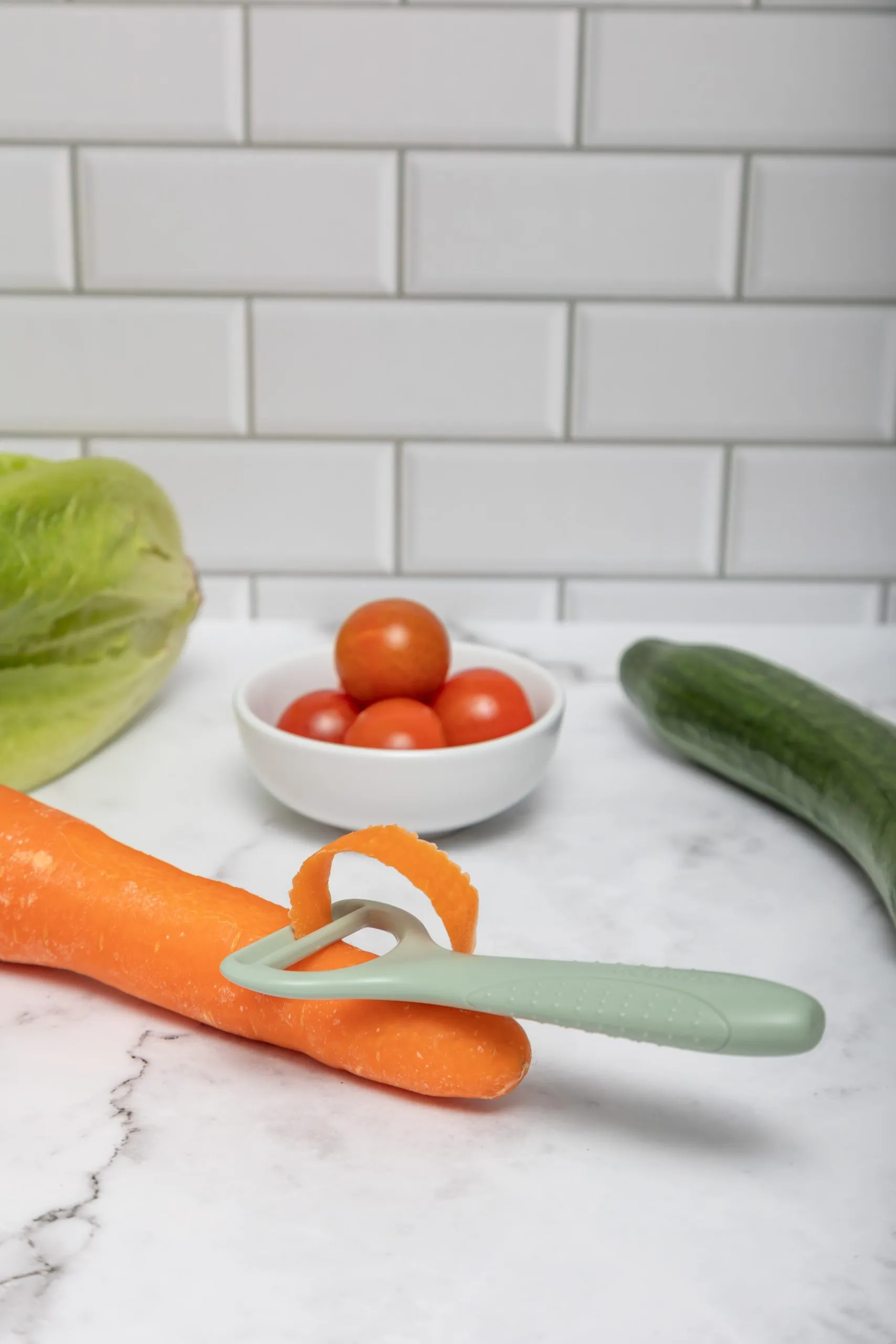 KiddiKutter Safer Peeler peeling a carrot beside tomatoes and cucumber