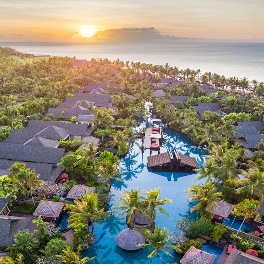 Aerial view of a tropical resort with lush greenery, multiple villas, and a large lagoon-style pool at sunset overlooking the sea—perfect for those seeking exclusive luxury tour packages.