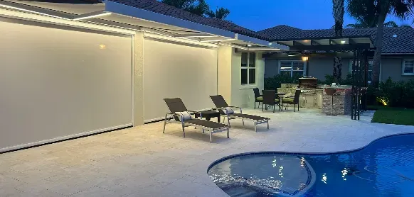 Modern backyard at dusk with two lounge chairs on a tiled patio next to a pool with a small round spa. A covered outdoor kitchen and grill area are visible in the background, surrounded by greenery.