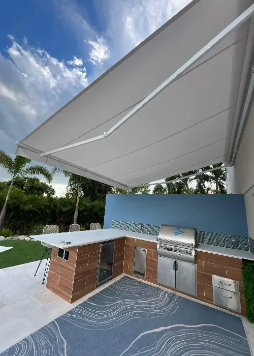 Modern outdoor kitchen with a large canopy, stainless steel grill, sink, and wood cabinetry. There&rsquo;s a blue backsplash and a patterned rug, surrounded by palm trees and greenery under a partly cloudy sky.