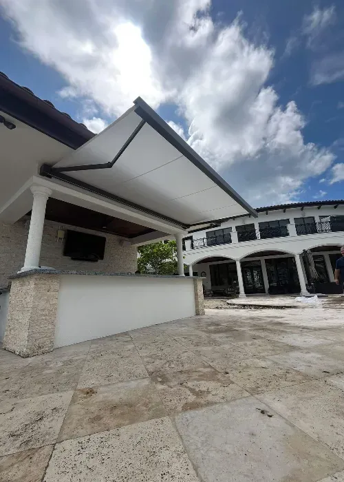 Outdoor bar area with a stone counter and extended white awning, in front of a large, two-story house with columns and balconies under a partly cloudy sky.