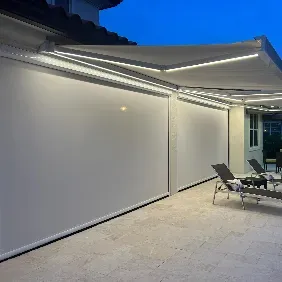 A modern patio at dusk features retractable lighted awnings and closed white screen shades. Two lounge chairs and a table sit on a tiled floor. The setting is illuminated by built-in LED strip lights.