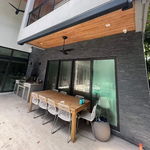 A modern outdoor patio with a wooden dining table, six white woven chairs, a built-in stainless steel grill, and large glass doors against a dark stone wall under a wooden overhang.