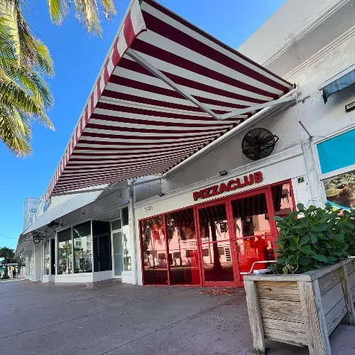 A sidewalk view of a white building with red-framed windows and a red-and-white striped awning reading "Pizza Club." Palm tree leaves and a planter with greenery are visible in the foreground.