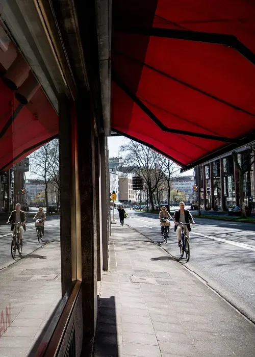 Two people ride bicycles down a city street under a large red awning. Their reflections appear in a shop window, and trees and buildings line the background on a sunny day.