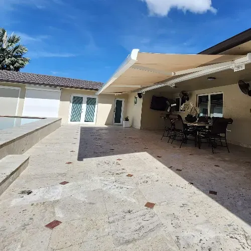 A spacious stone patio with a covered outdoor dining area, table and chairs, set beside a modern beige house and a pool under a bright blue sky.