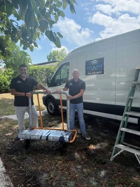 Two men stand next to a white van labeled "Miami" while holding a dolly with a large rolled item. They are outdoors, on grass near a leafy tree and a green ladder, smiling at the camera.