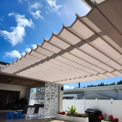 A modern patio with a retractable beige fabric canopy providing shade, outdoor seating, potted plants, and a white decorative privacy screen under a bright blue sky with scattered clouds.