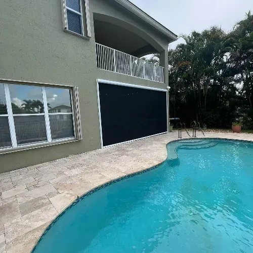 A backyard with a curved swimming pool, stone tile deck, and a two-story house with large windows, a balcony, and a screened patio. Lush green trees line the background.