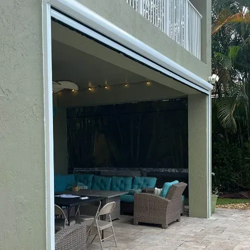 A covered outdoor patio with wicker furniture, turquoise cushions, a folding table, and string lights, surrounded by greenery and partially enclosed by a screen.