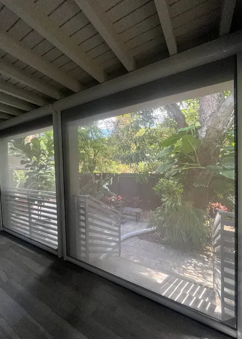 View through large glass windows with mesh screens, showing a lush garden with tropical plants, trees, and a metal gate on a sunny day. Indoors, the floor is wooden and the ceiling has exposed beams.
