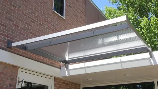 A modern metal and glass awning extends over a doorway attached to a brick building, providing shade and protection. Green trees and a clear blue sky are visible in the background.