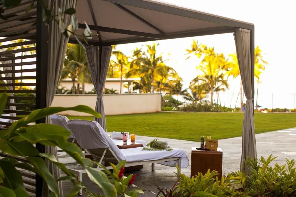 A shaded cabana with two lounge chairs sits on a stone patio overlooking a green lawn and palm trees. Drinks and snacks rest on a small table, creating a relaxing tropical outdoor setting.