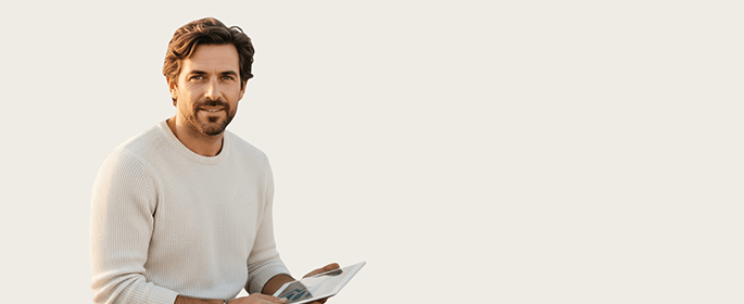 A man with brown hair and a beard, wearing a light sweater, sits holding a tablet. He is facing slightly left and smiling gently. The background is plain and light-colored, creating a minimalistic look.