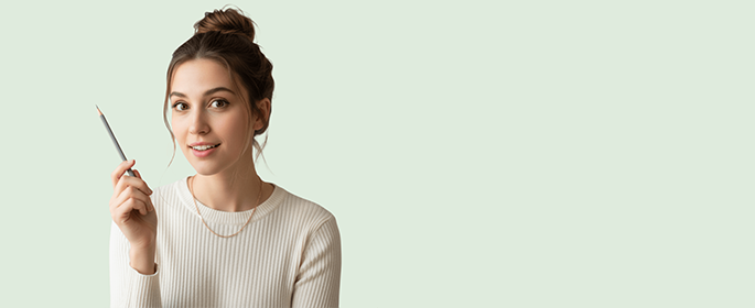 A young woman with brown hair in a bun, wearing a cream ribbed sweater and a gold chain necklace, holds a pencil up with a slight smile. She is facing forward against a light mint green background. The right side of the image is empty.