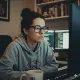 A person with dark hair in a bun, wearing glasses and a gray hoodie, sits at a desk using a computer. Bookshelves and two monitors are visible in the background. A white mug is on the desk in front of them. The room appears to be a home office.