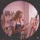 A woman with long blonde hair, wearing a dark apron over a white shirt, is painting on a canvas in a bright, art-filled studio. Shelves with art supplies are in the background. The image has a circular, slightly pinkish filter.