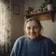 An elderly woman with gray hair tied back sits indoors, wearing a blue sweater and smiling gently. Behind her, a wooden shelf holds teapots and knick-knacks. Light streams through a window with patterned curtains, creating a warm, cozy atmosphere.