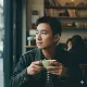 A young man with short dark hair sits by a window in a cozy café, holding a cup with both hands. He looks thoughtfully outside. Shelves with books and decorations are visible in the softly lit background, along with another person sitting behind him.