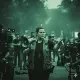 A person stands in the middle of a crowded street, raising their arms as police officers with shields and helmets close in from both sides. The scene is tense, with dark green tones and a sense of confrontation or protest.