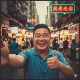 A smiling man in a blue polo shirt holds a phone for a selfie on a bustling street market at dusk, giving a thumbs-up. Colorful market stalls and neon signs, including one with Chinese characters, fill the lively background.