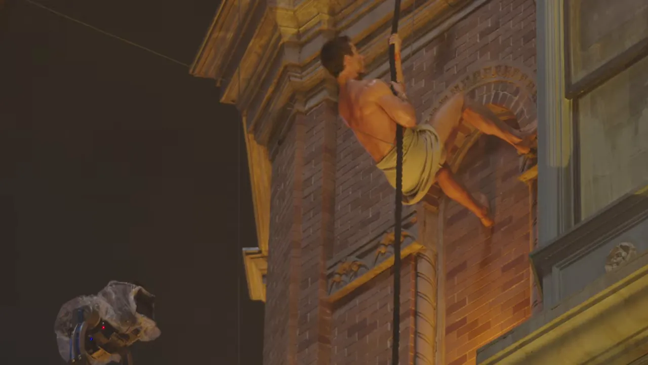 Actor in a towel climbing the exterior of a brick building on a rope at night