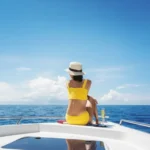 A woman in a yellow swimsuit and wide-brimmed hat sits on the deck of a white boat, gazing at the blue Nantucket ocean. She looks relaxed under the bright sun, glass beside her, savoring a Five-Star Weekend by Elin Hilderbrand.
