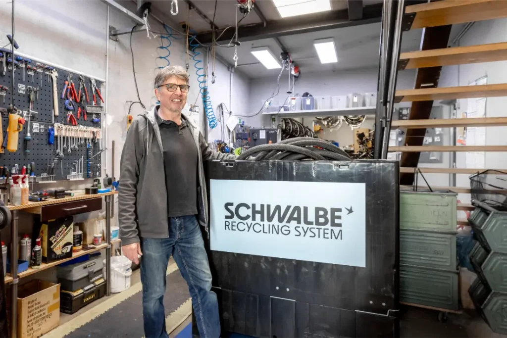 Un hombre junto a una máquina rotulada "Schwalbe Recycling System" en un taller, donde se ven herramientas y equipos que ponen de relieve el compromiso de Schwalbe con el reciclaje de neumáticos.