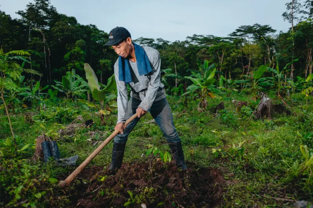 Una persona con gorra, botas y manga larga utiliza una azada para trabajar la tierra cerca de Schwalbe neumáticos en un verde campo agrícola tropical rodeado de árboles.