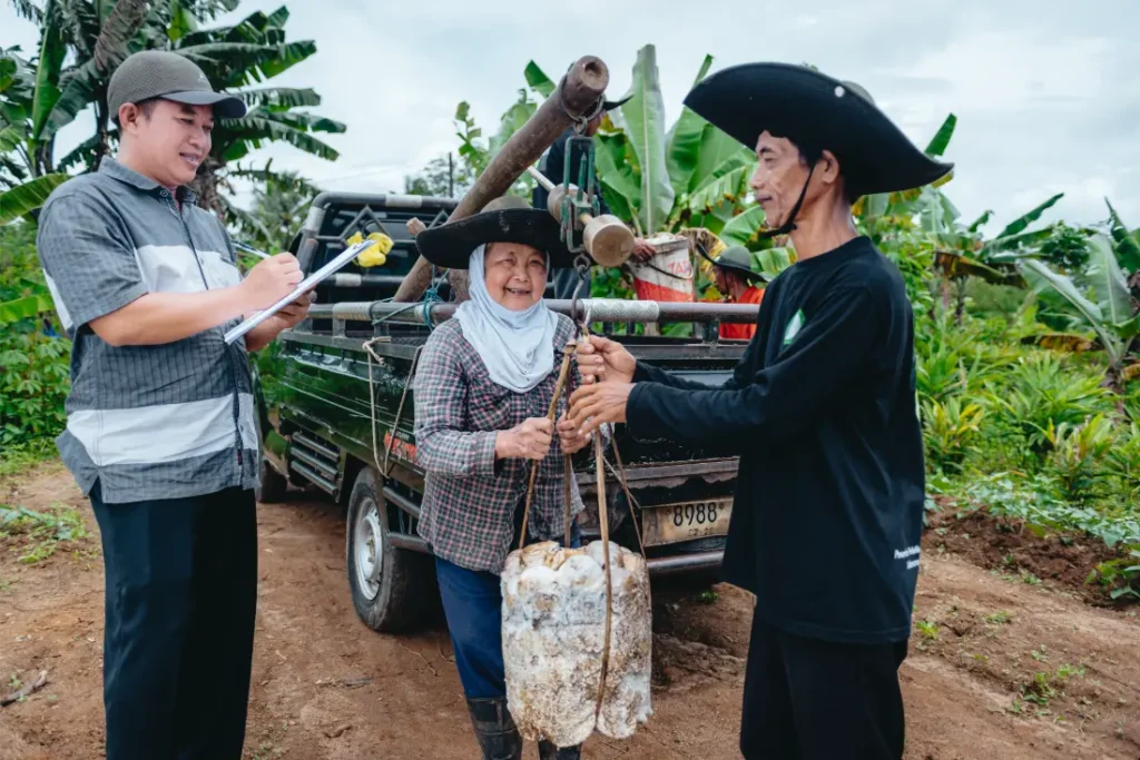 Tres personas permanecen junto a una camioneta con neumáticos Schwalbe en un camino de tierra; una escribe en un portapapeles mientras las otras dos manipulan un gran contenedor, rodeado de plantas tropicales.