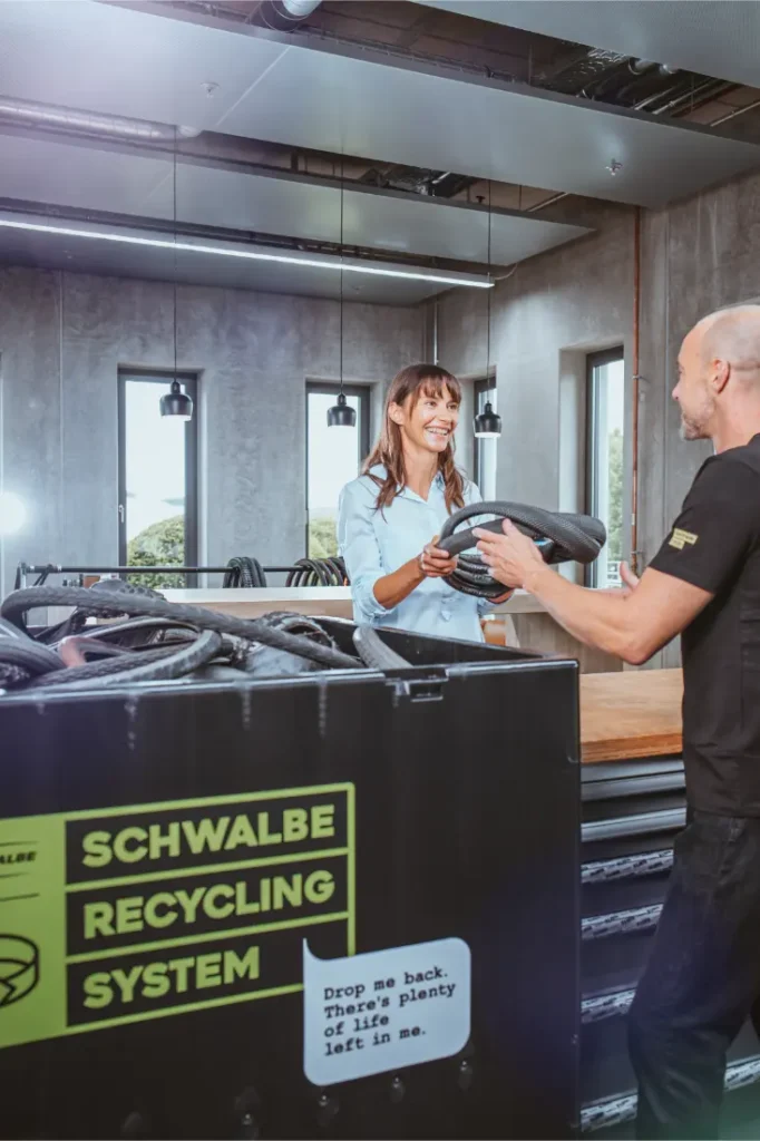 Dos personas junto a un contenedor del Sistema de Reciclaje Schwalbe, intercambiando neumáticos Schwalbe usados en un moderno ambiente interior.