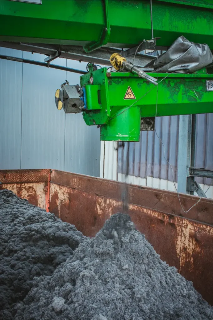 Una máquina industrial verde dispensa material fibroso gris en un gran contenedor metálico en el interior de una fábrica, parte del proceso de producción de neumáticos Schwalbe.