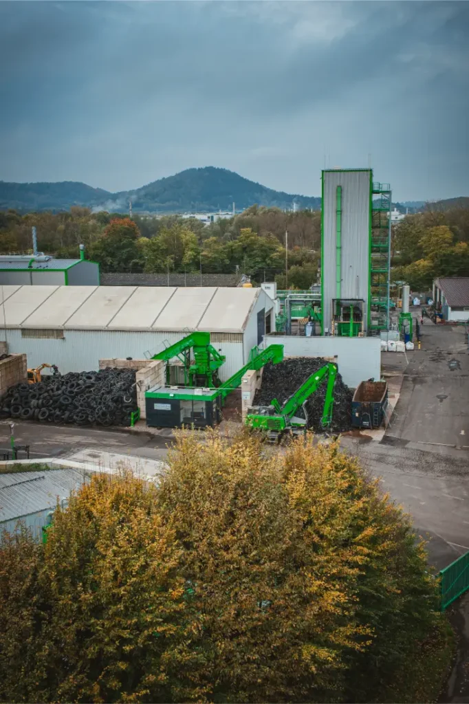 Una instalación de reciclaje con maquinaria Schwalbe verde moviendo pilas de neumáticos, rodeada de árboles y colinas bajo un cielo nublado.