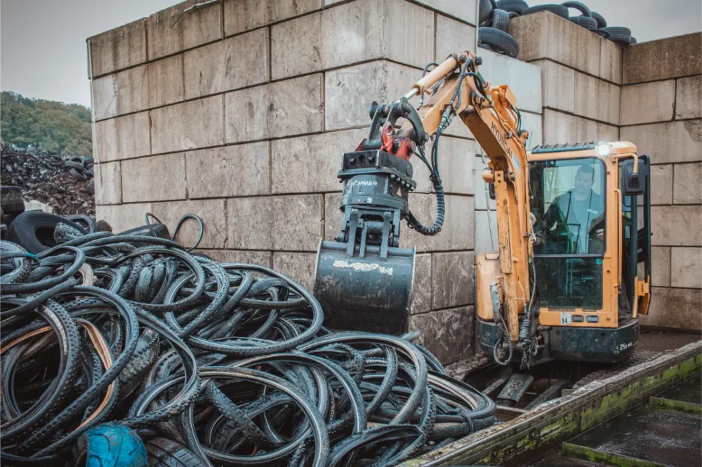 Una excavadora con garra levanta una pila de neumáticos usados en una planta de reciclaje industrial.
