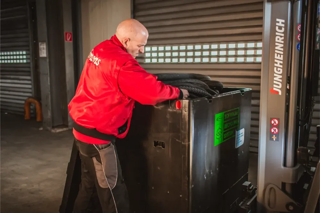 Una persona con chaqueta roja mueve o inspecciona un gran contenedor industrial negro junto a una carretilla elevadora Jungheinrich con neumáticos Schwalbe en el interior de un almacén.