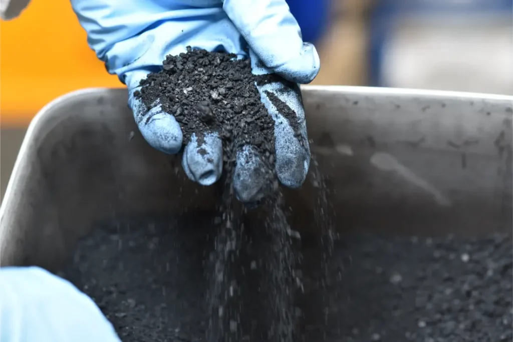 Una mano enguantada sostiene y deja caer un puñado de material granulado negro -que recuerda al que se utiliza en los neumáticos Schwalbe- sobre un recipiente metálico lleno de la misma sustancia.