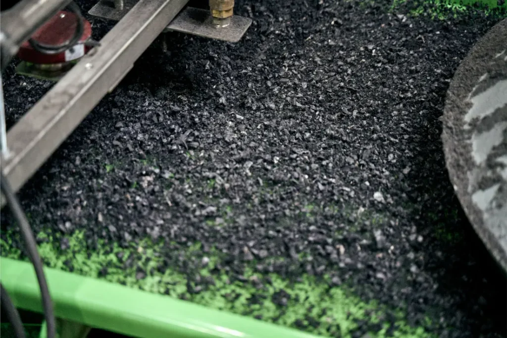 Primer plano de una máquina que procesa pequeños gránulos de caucho negro a partir de neumáticos reciclados en una cinta transportadora verde en un entorno industrial, mostrando el compromiso de Schwalbe con las prácticas sostenibles.