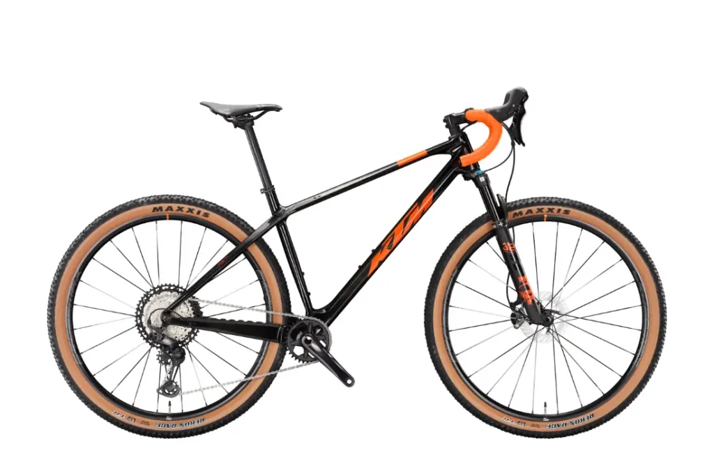 Se muestra una bicicleta de montaña negra y naranja, con manillar curvo y neumáticos de flanco marrón claro, en una vista lateral sobre un fondo blanco.