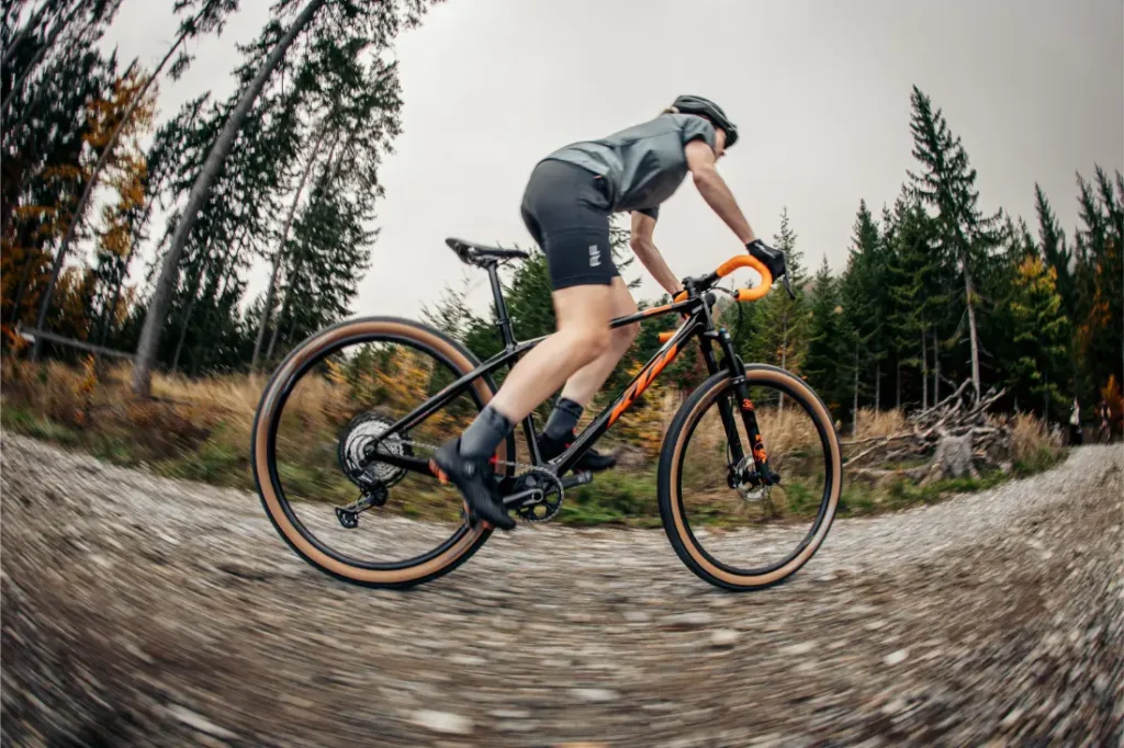 Una persona va en bicicleta de gravel por un sendero forestal, con casco y ropa de ciclismo. Al fondo se ven pinos y un cielo nublado.
