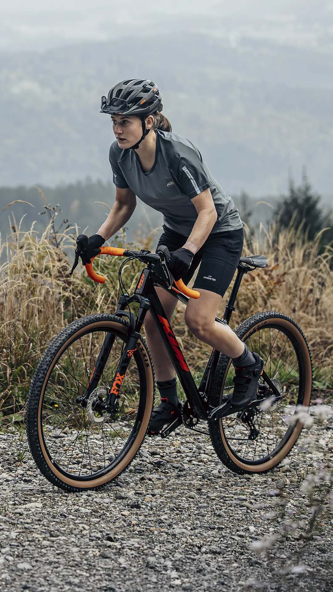 Una persona que lleva casco y equipación de ciclismo circula en una bicicleta de gravel por un sendero rocoso, con hierba alta y colinas al fondo.
