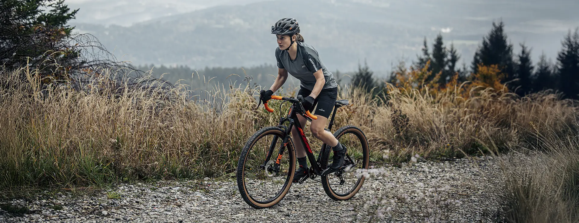 Un ciclista, que lleva casco y ropa de ciclismo, circula en una bicicleta de gravel por un sendero rocoso entre la hierba alta, con colinas y árboles al fondo.