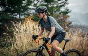 Una persona que lleva casco y ropa de ciclista va en bicicleta por un camino de tierra entre la hierba alta, con árboles al fondo.