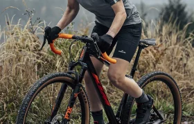 Una persona que lleva casco y equipación de ciclismo circula en una bicicleta de gravel por un sendero rocoso, con hierba alta y colinas al fondo.