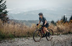 Una persona que lleva casco y equipación de ciclismo pedalea en una bicicleta de gravel cuesta arriba por un sendero rocoso, con un paisaje montañoso envuelto en niebla al fondo.