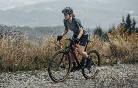 Un ciclista, que lleva casco y ropa de ciclismo, circula en una bicicleta de gravel por un sendero rocoso entre la hierba alta, con colinas y árboles al fondo.