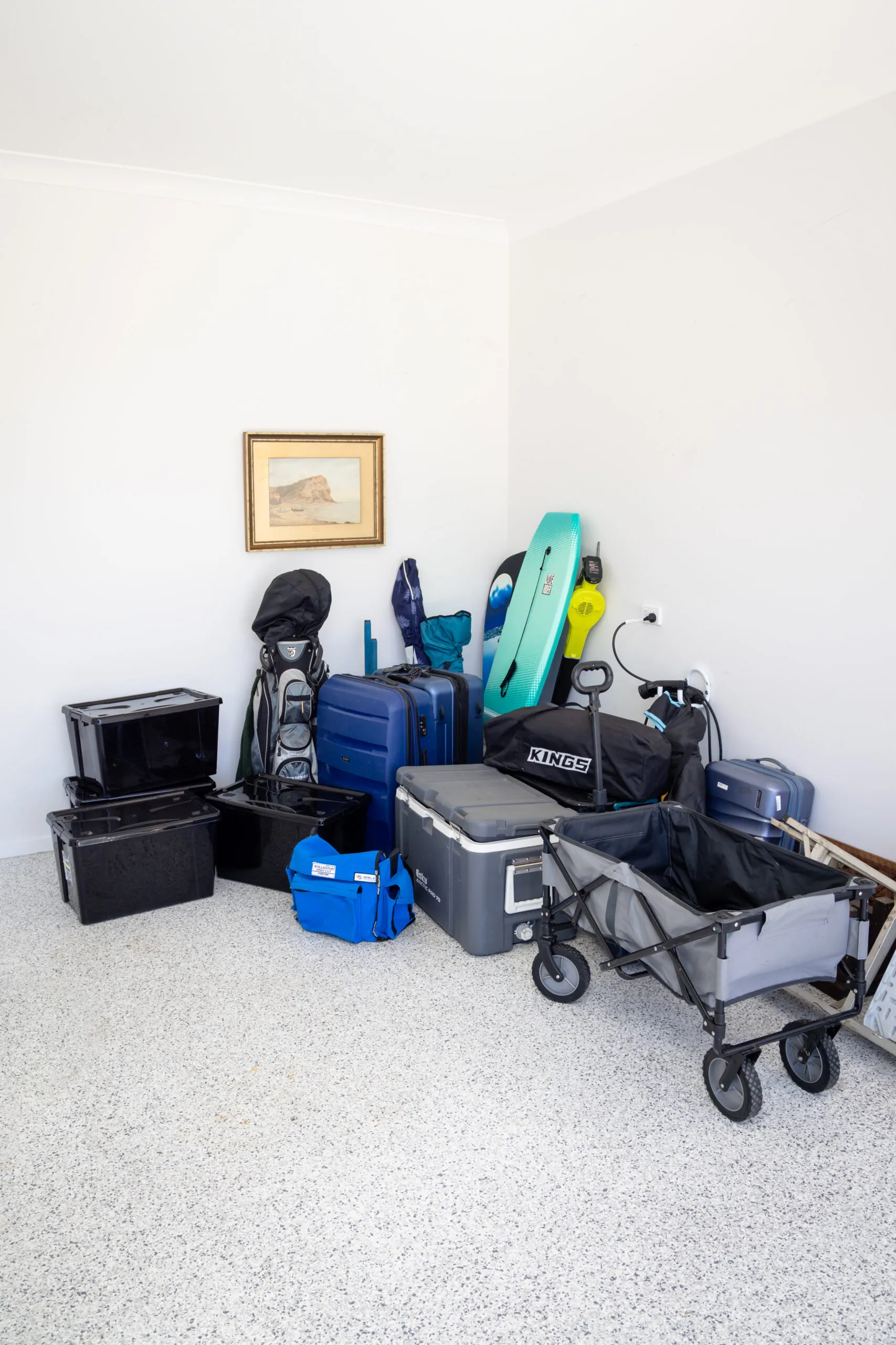Corner of a white-walled storage room with suitcases, a turquoise paddleboard, stroller, bags, and a framed landscape painting on the wall.