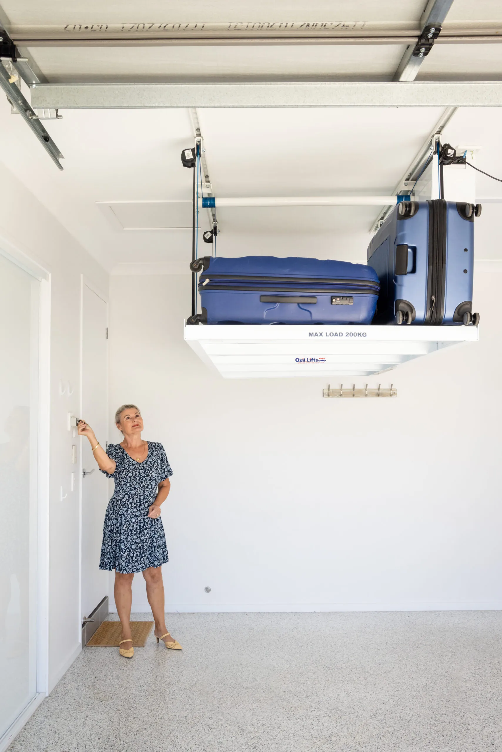 Woman in a blue floral dress reaching to operate a ceiling-mounted luggage hoist carrying two blue suitcases in a bright room.