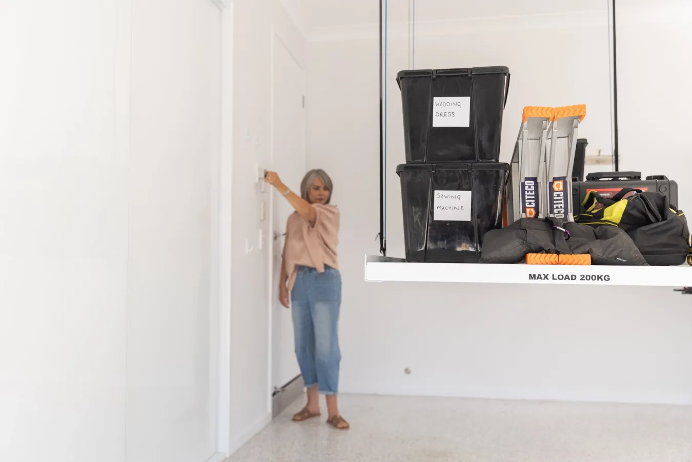 Woman standing near a doorway in a bright room, beside a storage platform with labeled boxes reading 'Wedding Dress' and 'Sewing Machine'.